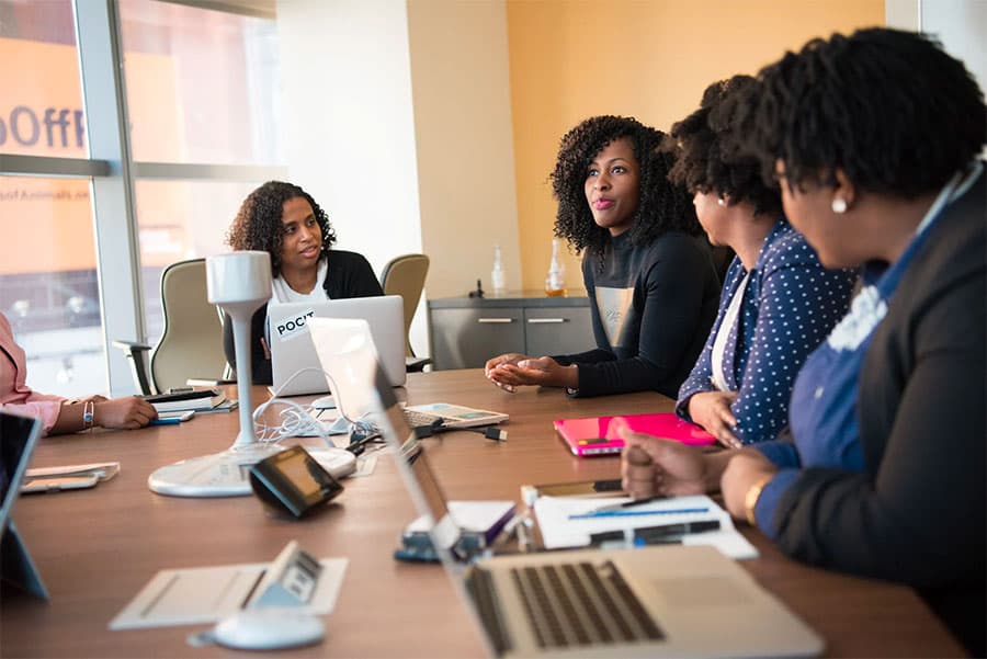 Profissionais reunidas em volta de uma mesa de reunião, com laptops e anotações, durante uma conversa de trabalho.